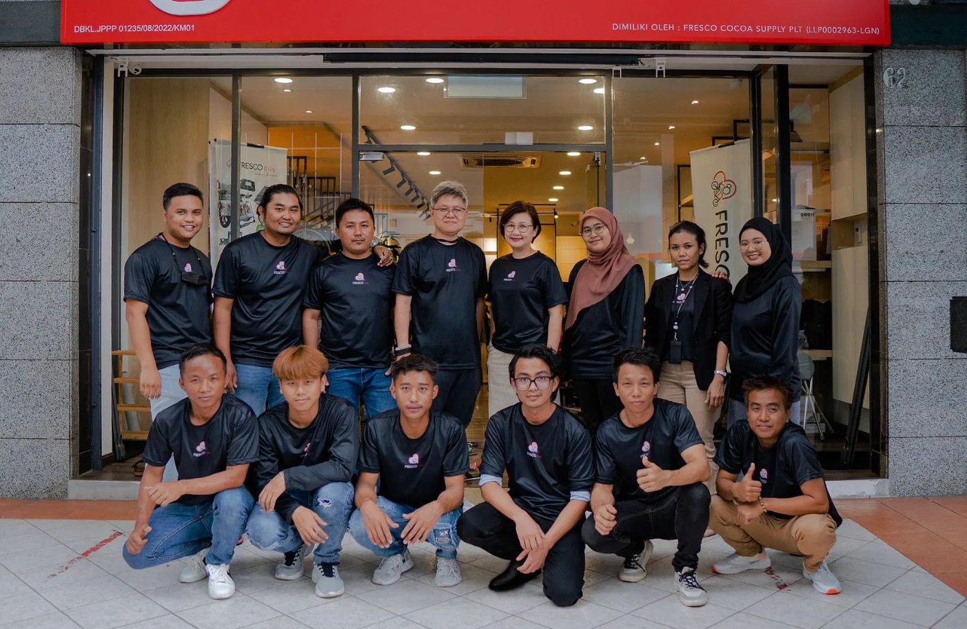 A group of fifteen people in matching black shirts pose for a photo in front of a storefront.