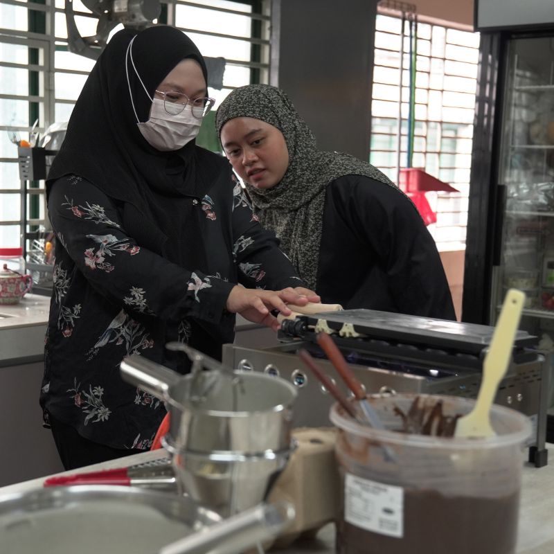 Two women in headscarves work together in a kitchen, using a griddle. One wears a face mask. Various kitchen tools and ingredients are on the counter.