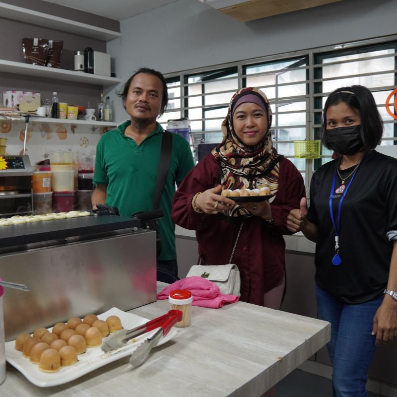 Three people standing in a kitchen, one holding a plate of food. Counter displays various items, including eggs. They are smiling, with one person wearing a mask.