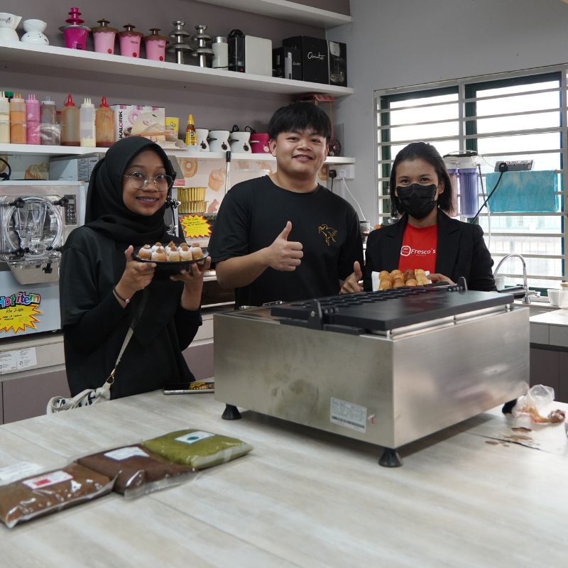 Three people standing in a kitchen with one holding a plate of food, next to a stainless steel appliance on the counter.