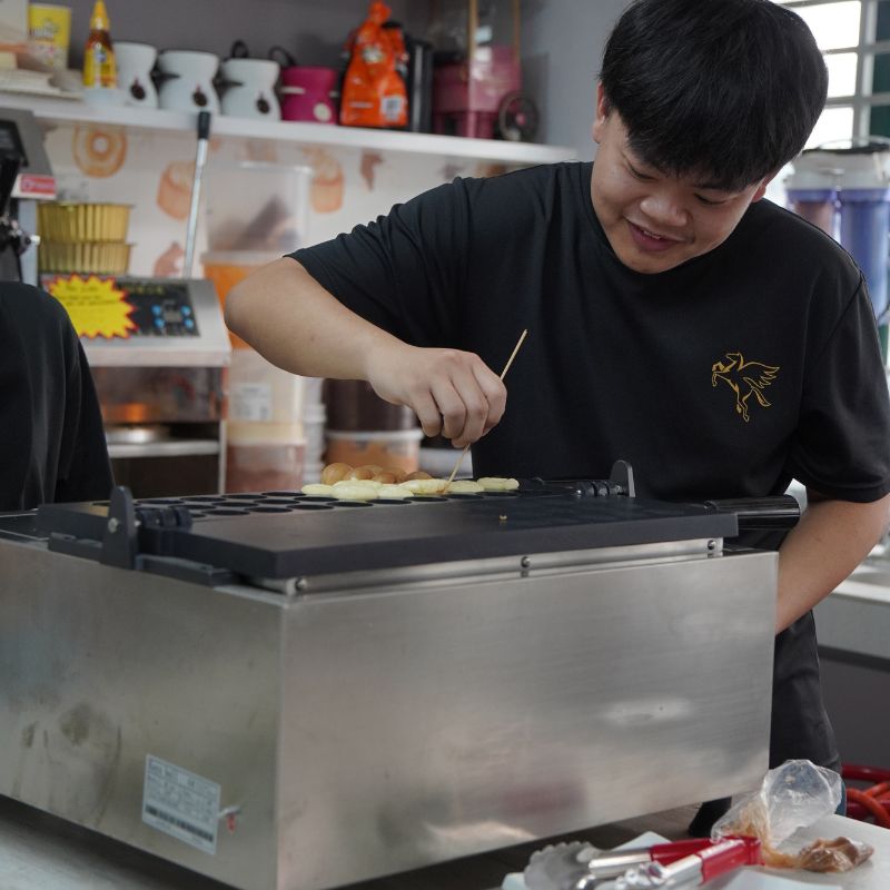 A person cooking round pastries on a griddle, using a skewer for flipping. Shelves with cooking supplies are in the background.