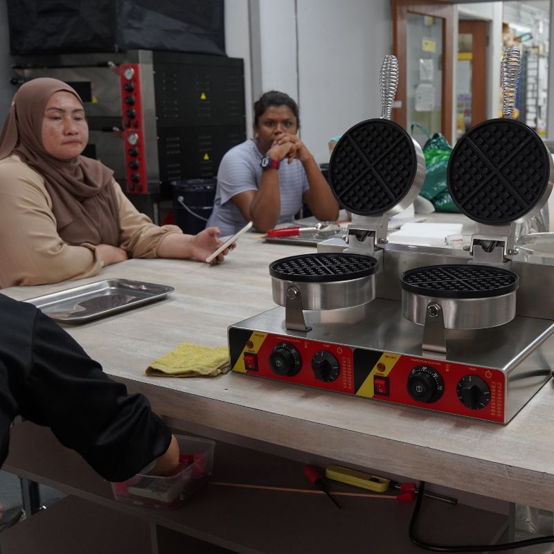 Two people sit at a table with waffle makers in a kitchen setting. One person holds a phone, while the other rests their chin on their hands.