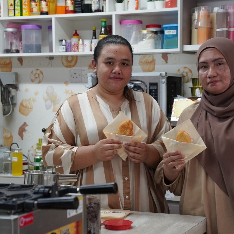 Two people in a kitchen holding pastries. Shelves with jars and bottles are visible in the background.
