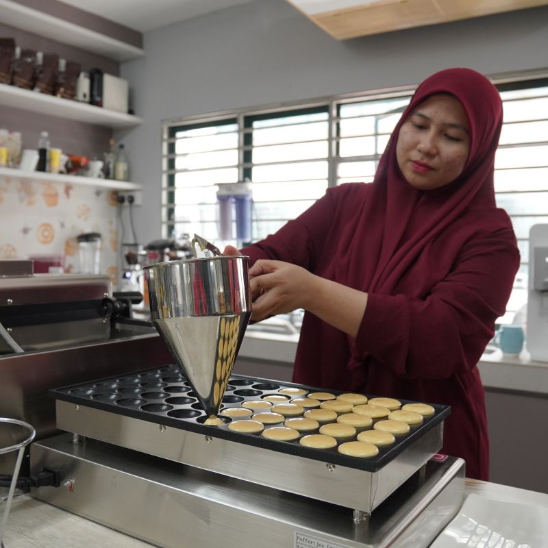 A woman in a maroon hijab pours batter into circular molds on a pan in a kitchen setting.