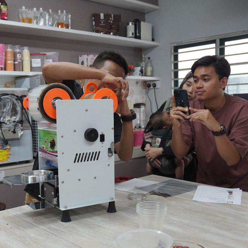 People observing a machine demonstration in a kitchen. One person records with a phone, while another adjusts the machine. Shelves with items are in the background.