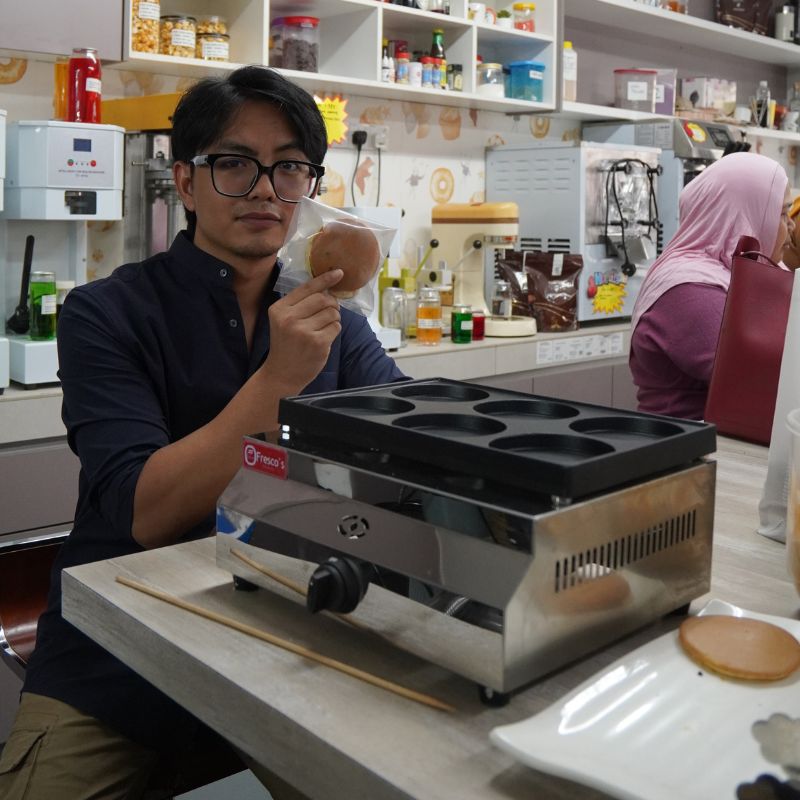 A person sits at a kitchen counter, holding a packaged item. A pancake griddle and a partially visible person in a pink headscarf are nearby. Shelves with various kitchen items are in the background.