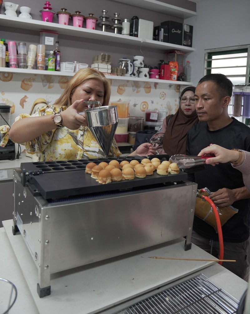 A woman pours batter into a bubble waffle maker, similar to a kayaball machine, while others watch in a kitchen filled with various tools and ingredients on shelves.