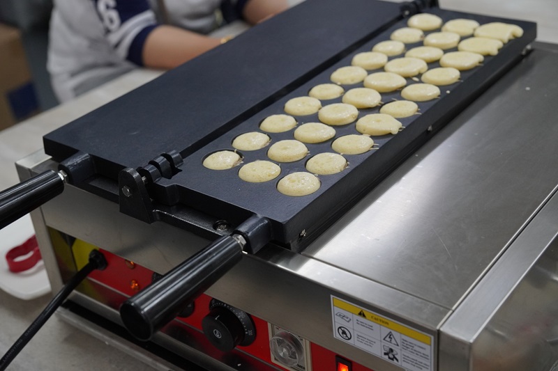 A kayaball machine griddle with small, round pancake batter portions cooking on it, with a person in the background.
