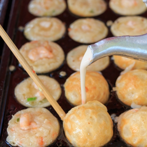 Batter is being poured into round molds on a griddle, making small savory pancakes with shrimp pieces, while a skewer is used to turn one pancake—just like in a takoyaki tutorial on how to make takoyaki at home.