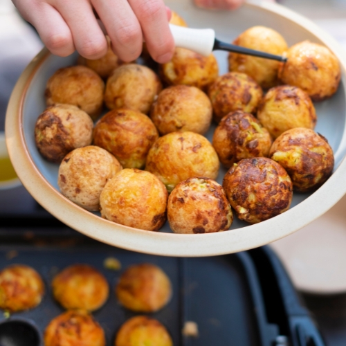 A hand holds a plate of golden brown takoyaki balls above a pan with more takoyaki, using a pick to lift one ball—just like in a takoyaki shop.