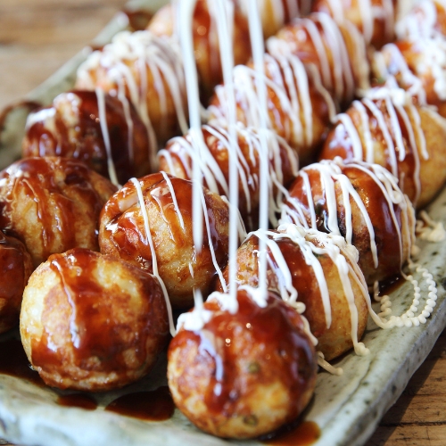 A rectangular plate holds rows of takoyaki balls topped with brown sauce and drizzled with white mayonnaise, just like you'd find at a takoyaki shop.