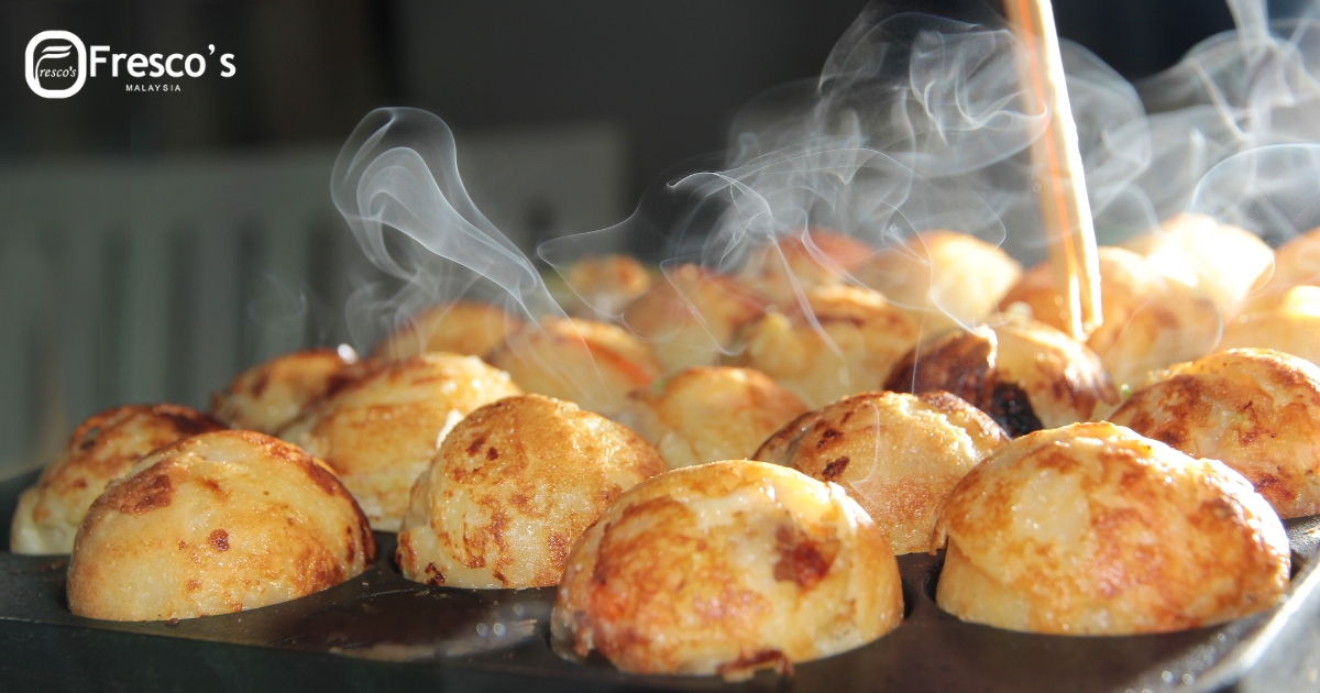 Close-up of golden-brown, steaming pastries in a baking tray, as a wooden skewer shapes one—perfect for a takoyaki tutorial or learning how to make takoyaki; "Fresco's Malaysia" logo in the corner.