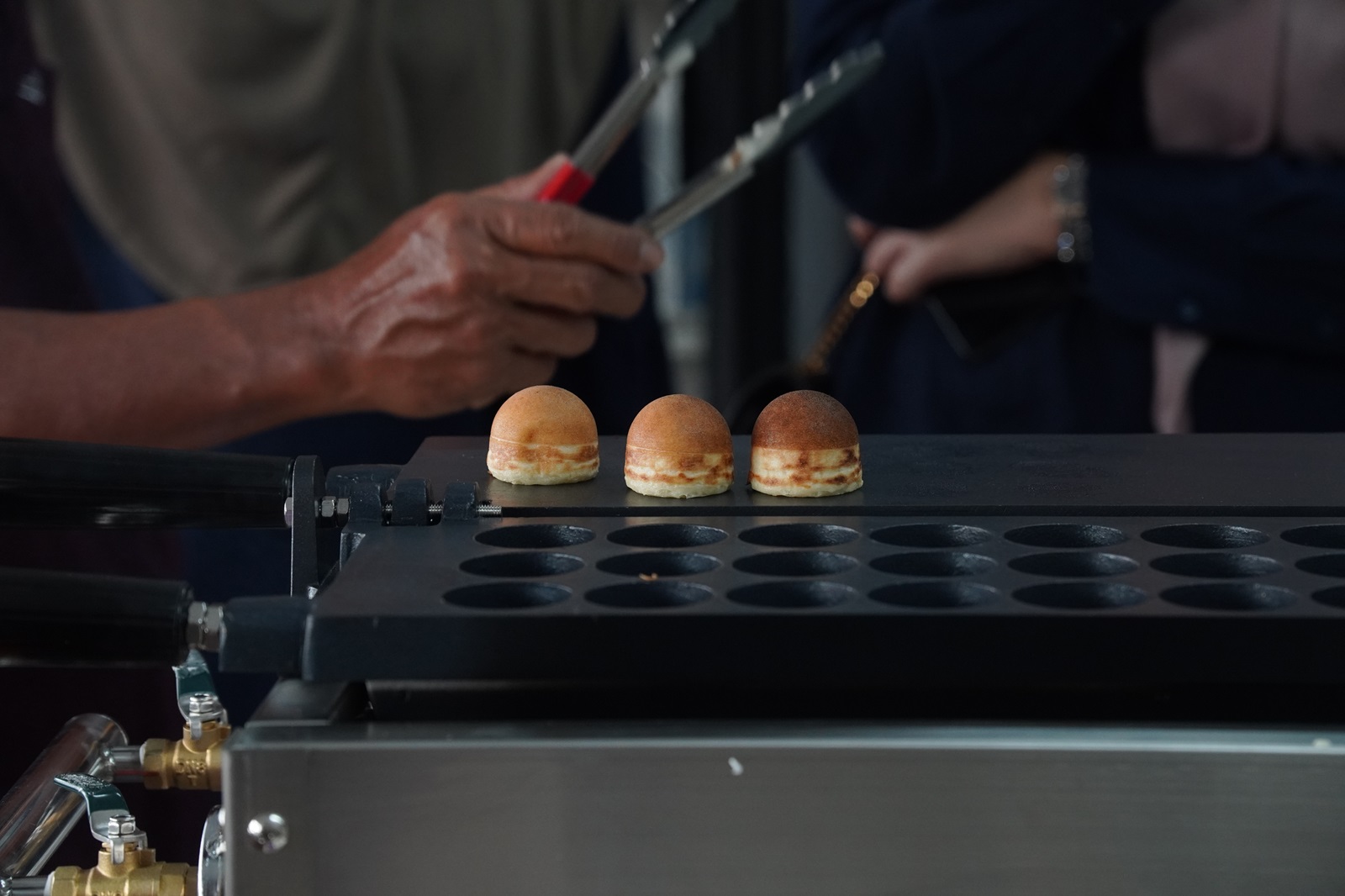 Three round pastries are cooking on a griddle with a kayaball machine, while a person holding tongs stands nearby.