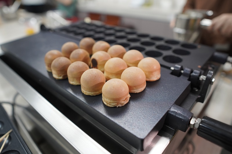 A row of round, golden-brown pastries from a popular kayaball shop sits on a mold pan, with more empty molds visible in the background on a kitchen counter.