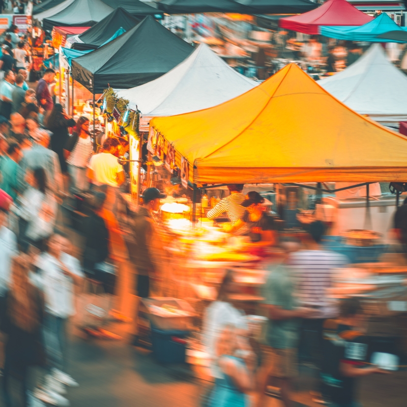 Blurred image of a busy outdoor market with colorful tents and crowds of people browsing stalls at dusk, including a Kayaball shop showcasing popular treats from kayaball Malaysia.