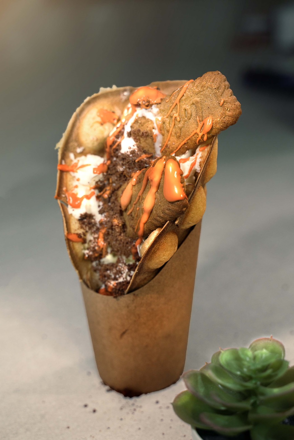A bubble waffle cone, made with a waffle and cone tool, is filled with ice cream, chocolate crumbs, a cookie, and orange sauce in a brown paper cup. A small succulent adds charm in the foreground.