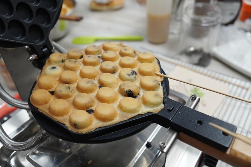 A bubble waffle is being cooked in a double function waffle machine, with some of the batter turning golden brown and slightly puffed up in each compartment.