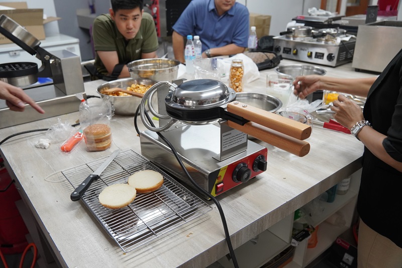 People prepare food around a countertop with a Double function waffle machine, sliced bread, kitchen utensils, and various ingredients.