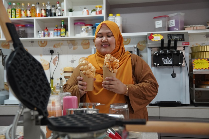 A woman in a kitchen holds two bubble waffles in cones, standing before shelves with bottles and equipment—she's showcasing treats made with a double function waffle machine.
