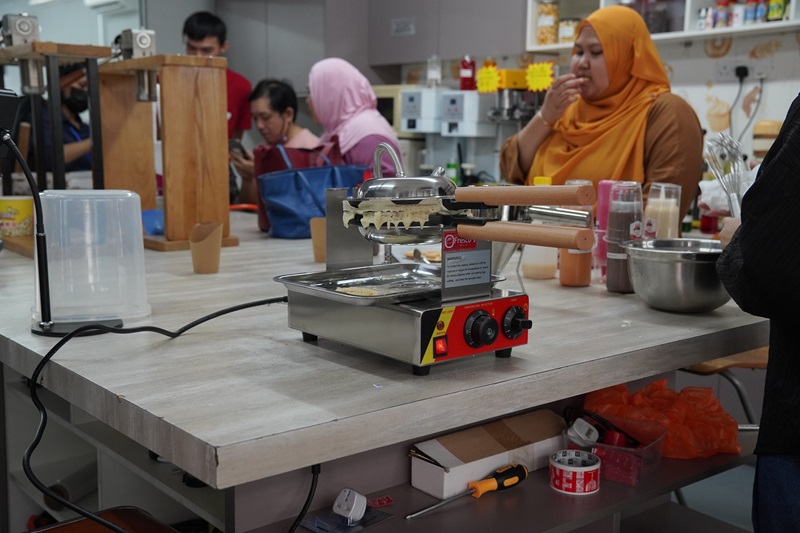 A double function waffle machine sits on a kitchen counter while several people, some in hijabs, work and talk in the background. Various kitchen tools and supplies are visible on the counter.