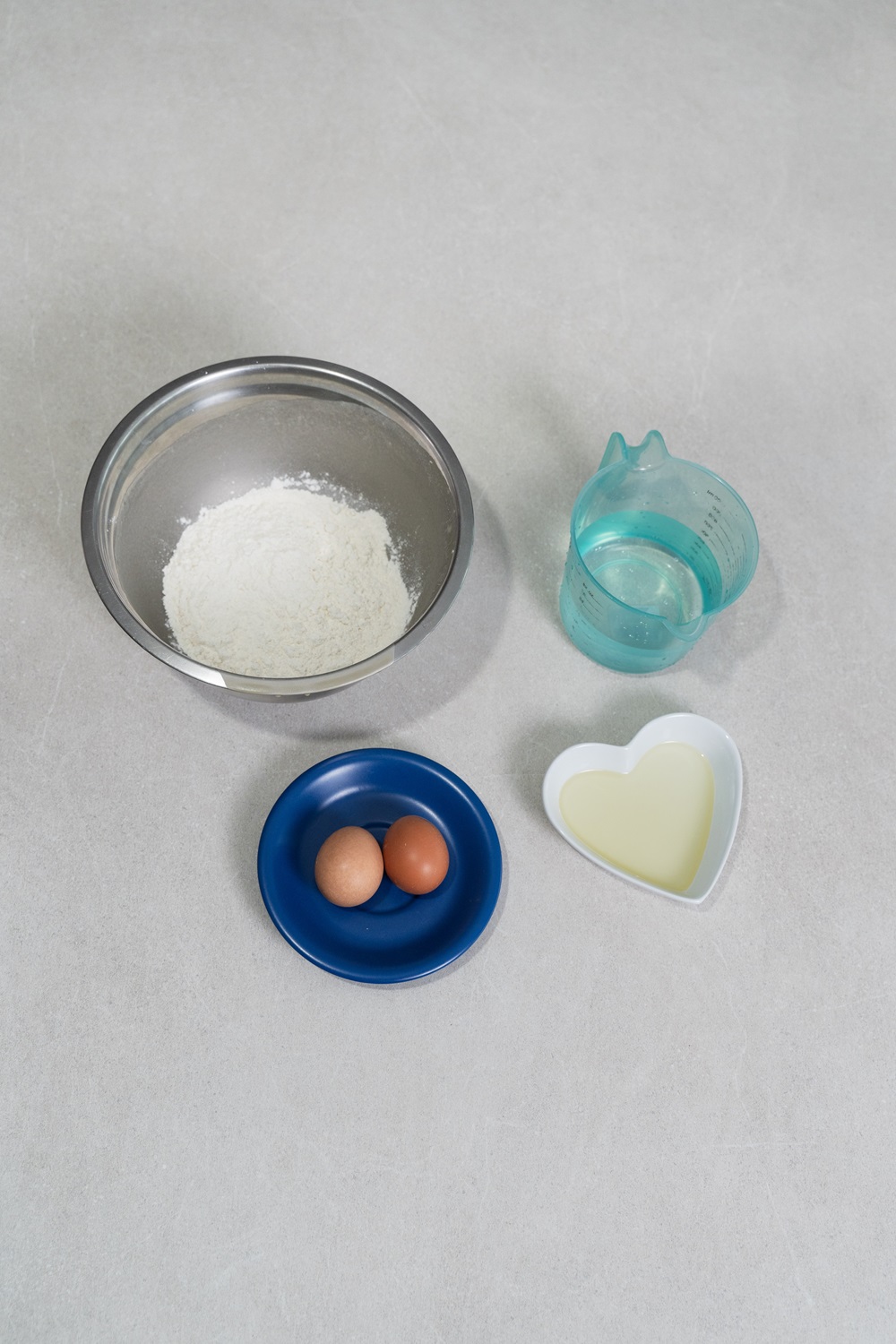 A metal bowl with flour, a blue plate with two eggs, a heart-shaped dish with oil, and a measuring cup with water are arranged on a light surface—perfect ingredients to use with your Egg Shape Waffle Maker.