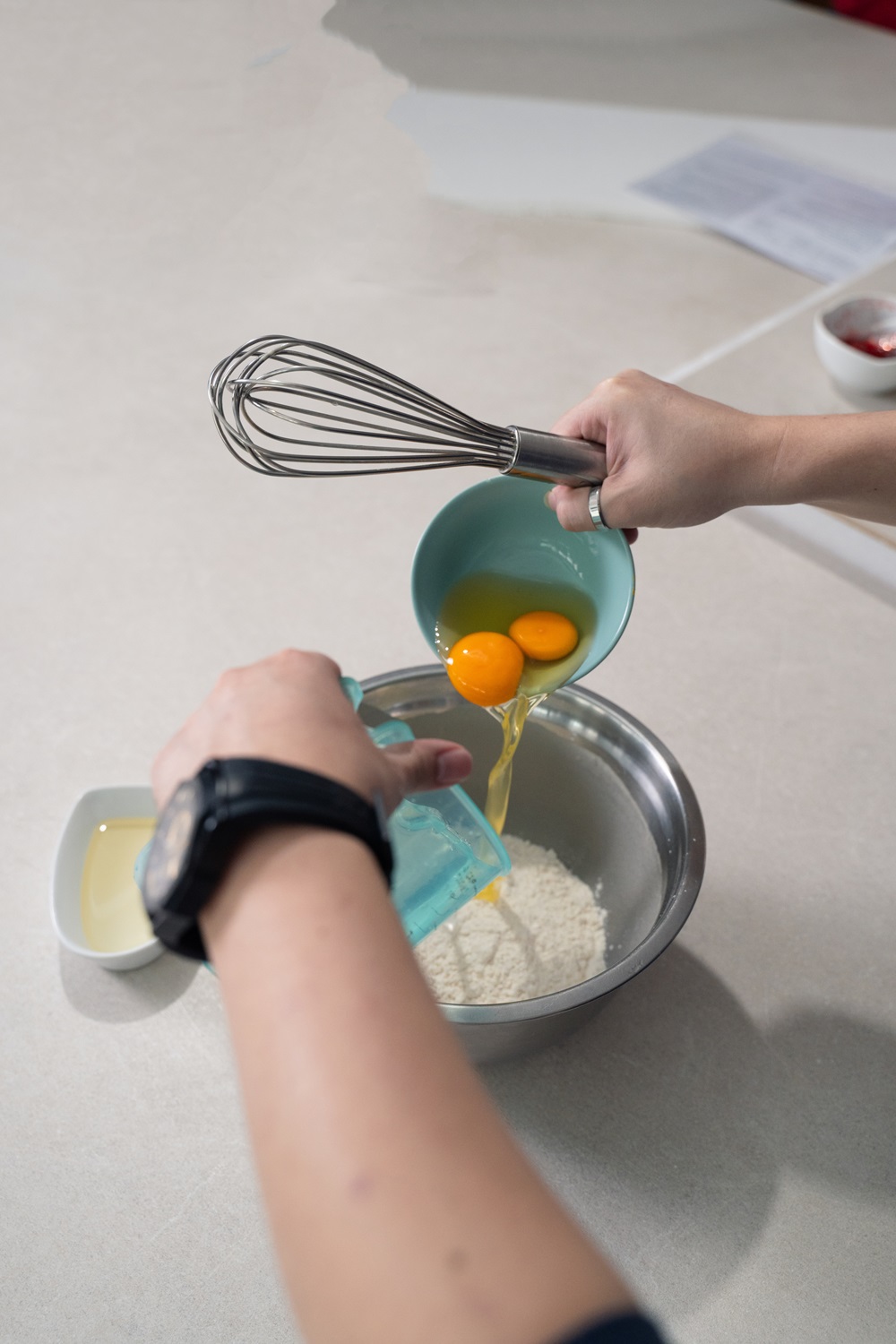 Two eggs are poured from a bowl into flour, while another person adds liquid from a measuring cup—perfect prep for creating treats with your Egg Waffle Machine and starting a small food business.