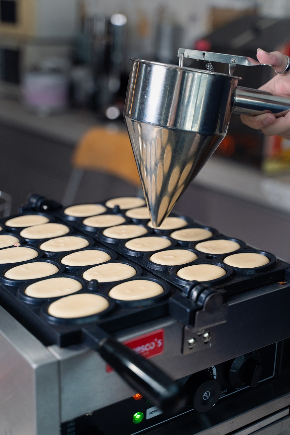 A person fills a circular mold pan with batter using a metal dispenser in a kitchen, perfect for making treats with an Egg Waffle Machine—an ideal way to Start Small Food Business ventures.