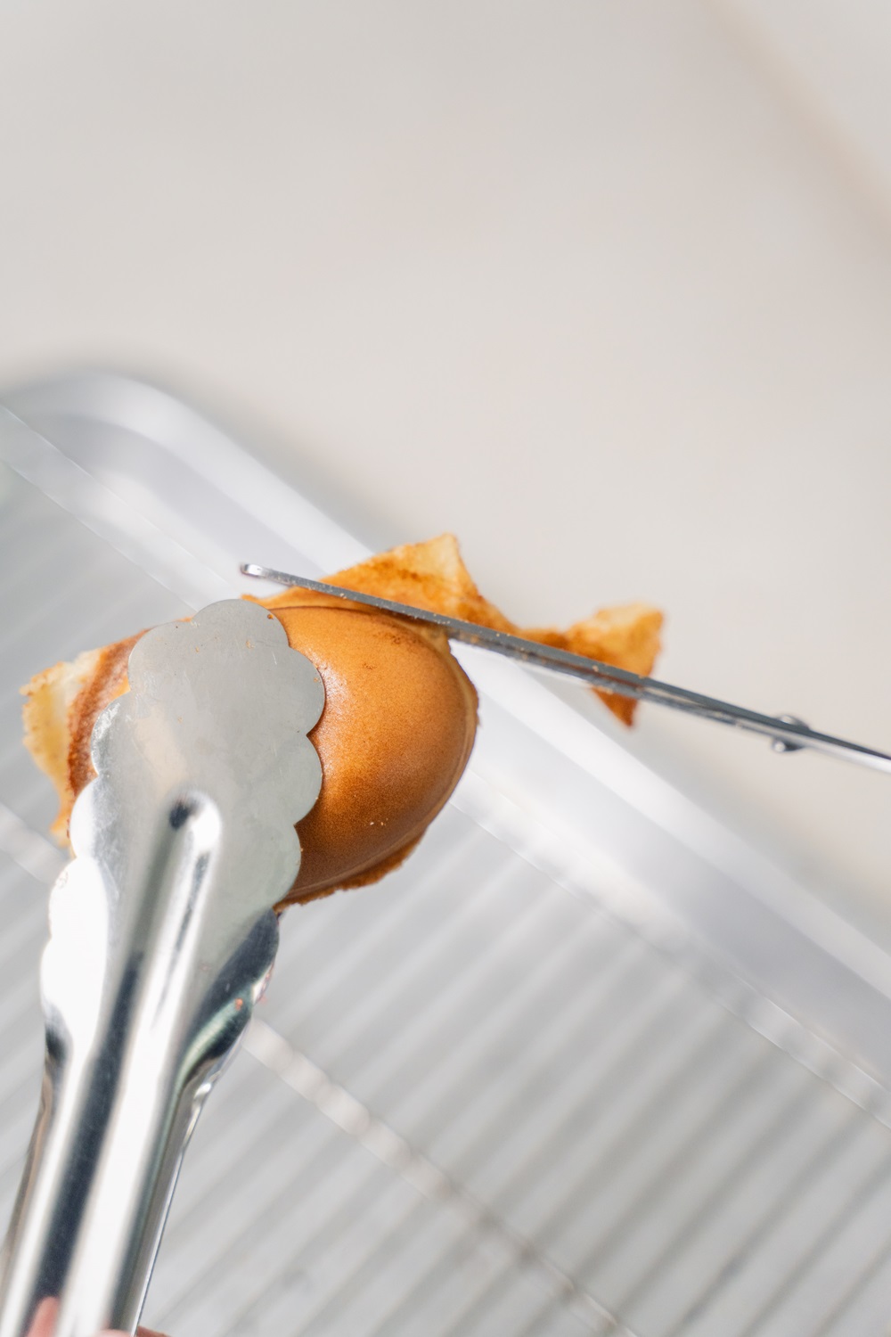 A pair of metal tongs holds a brown egg while a wire tool cracks the egg over a metal cooling rack—just the first step in using an Egg Shape Waffle Maker to kickstart your own profitable snack business.