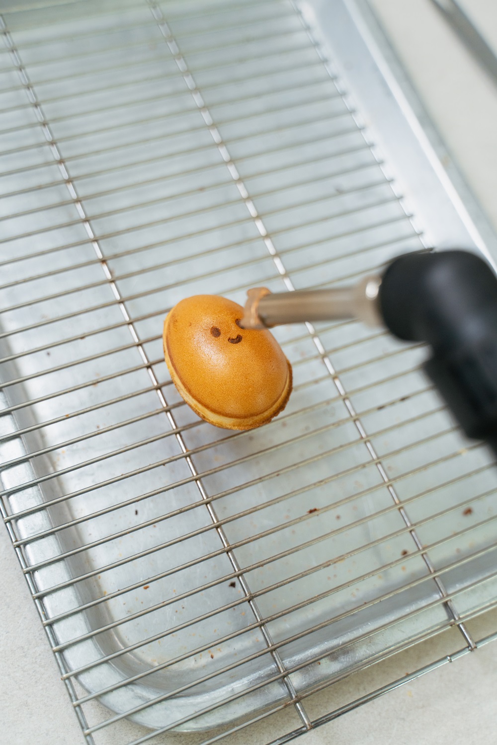 A blowtorch is being used to toast the surface of a small round pastry with a smiling face, resting on a wire cooling rack—perfect inspiration for anyone looking to start a small food business.