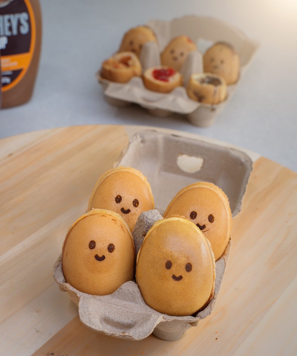 Four round pastries with smiling faces are placed in a small egg carton, showcasing how an Egg Waffle Machine can help create treats perfect for a Profitable Snack Business. More pastries are visible in the background.