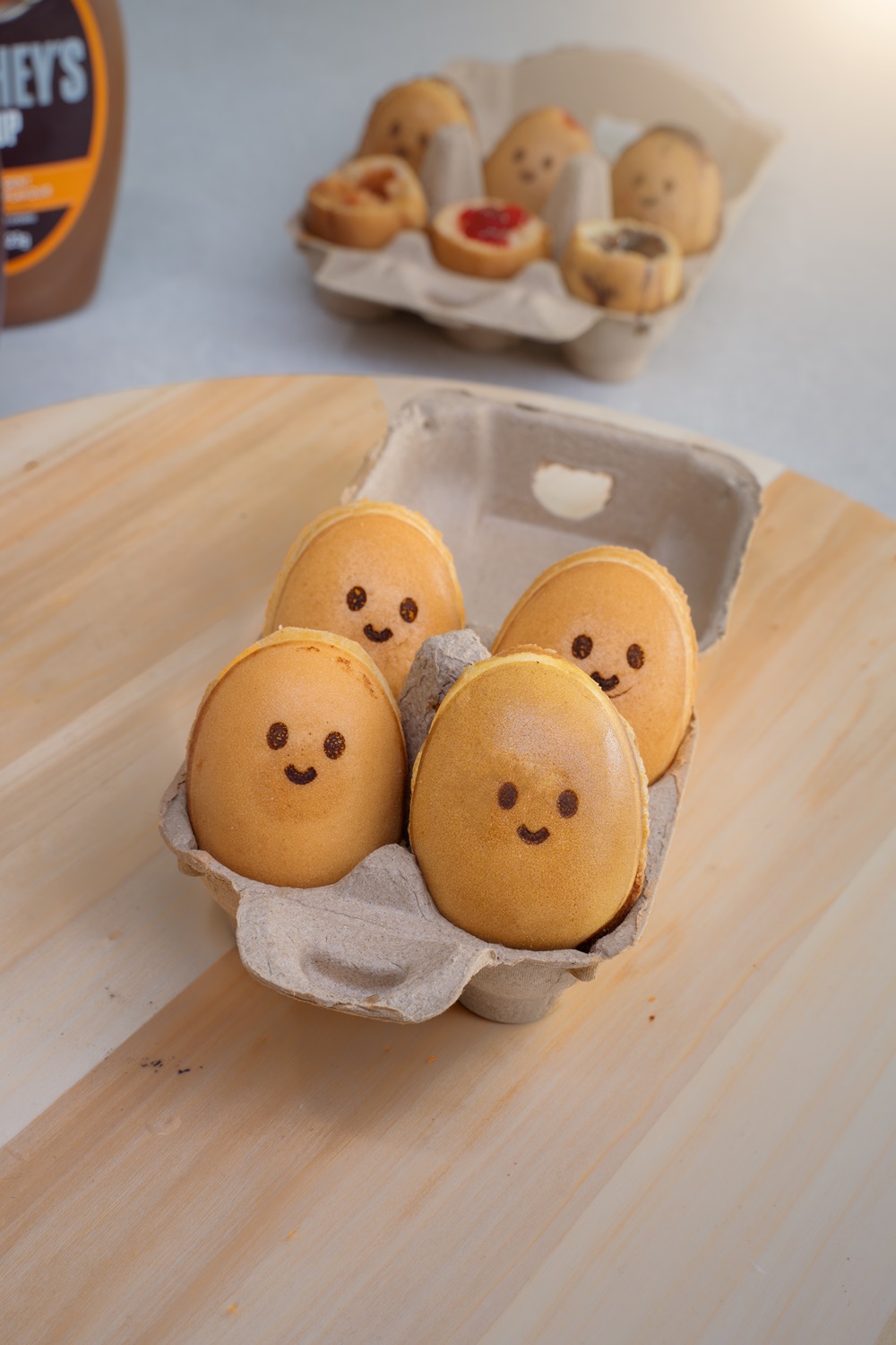 Four round pastries with smiling faces are placed in an egg carton, showcasing the creativity possible when you start a small food business using tools like the Fresco Malaysia Waffle Machine. More decorated pastries sit in another carton on a wooden surface.