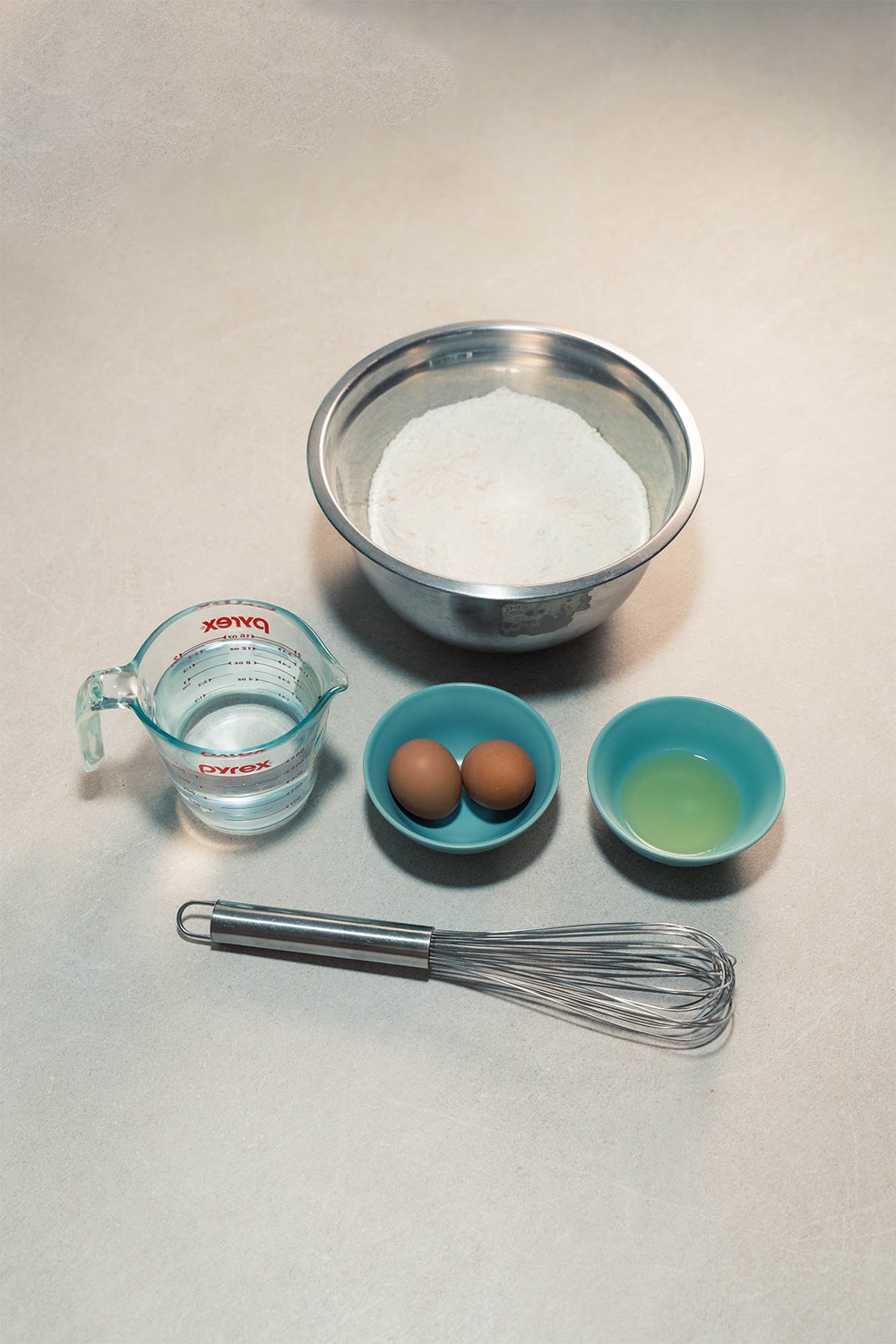 A metal bowl of flour, a measuring cup of water, two eggs in a small bowl, a bowl of lemon juice, and a whisk are arranged on the countertop—perfect for learning how to make waffle treats or testing your popsicle waffle machine Malaysia.