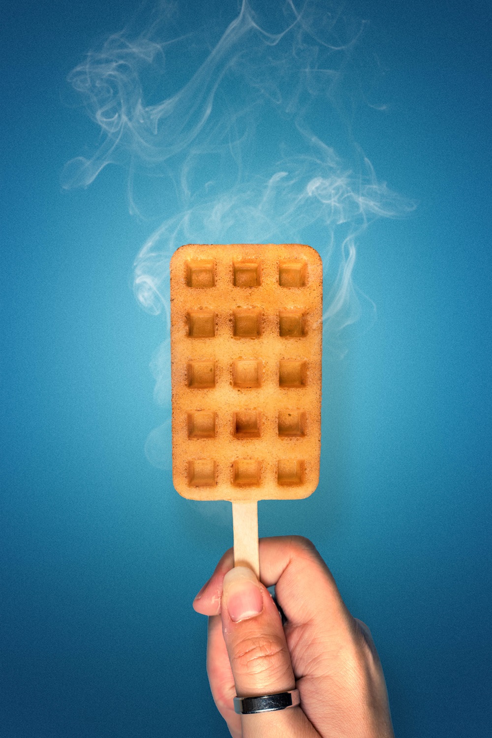 A hand holds a crispy popsicle waffle on a wooden stick against a blue background, with steam rising from the waffle—perfectly made using a Popsicle waffle machine Malaysia.