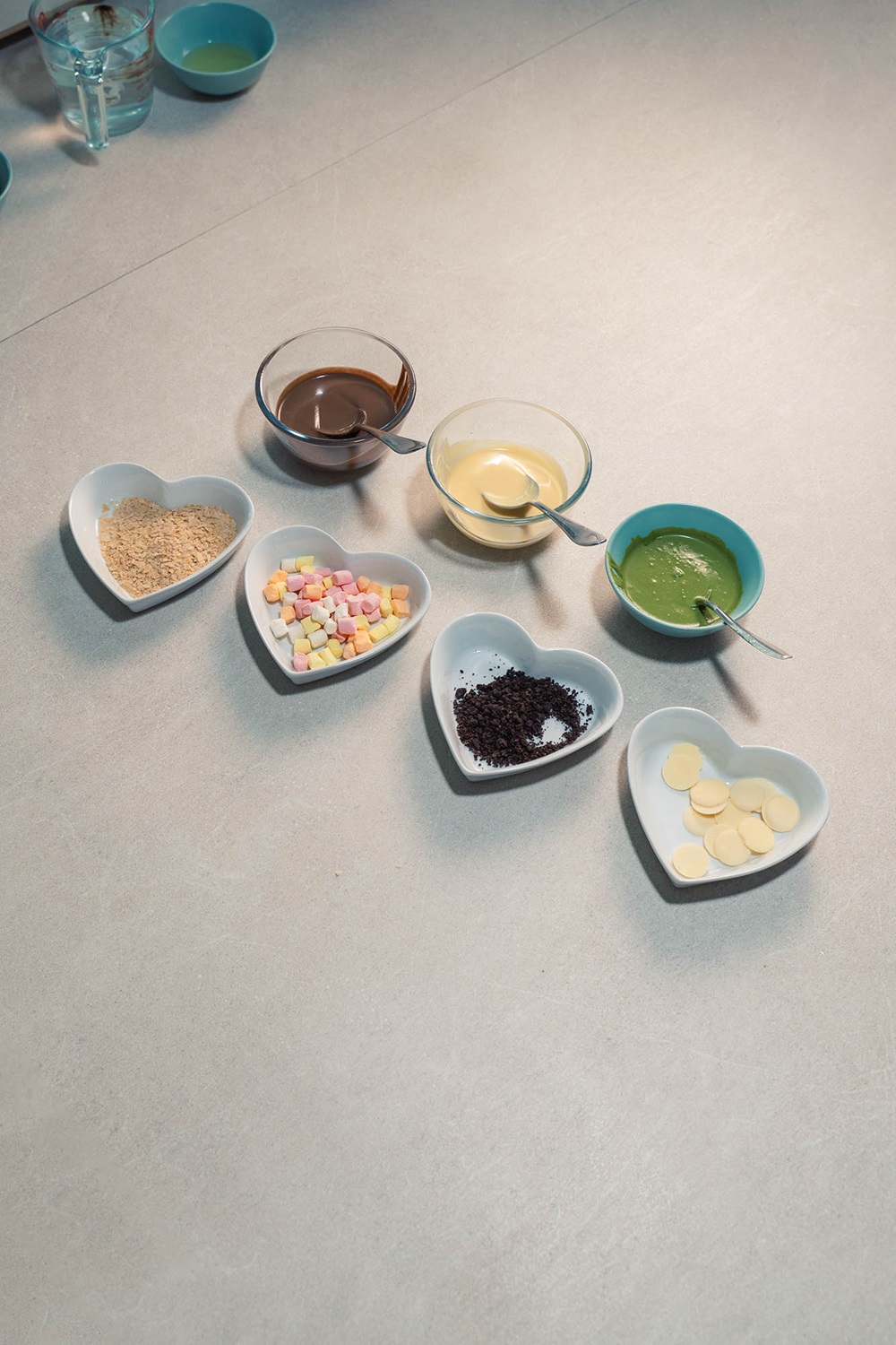 Six heart-shaped bowls contain graham crumbs, mini marshmallows, crushed cookies, and white chocolate buttons; three glass bowls hold melted chocolate, cream, and green sauce—perfect toppings for crispy popsicle waffles made with a popsicle waffle machine Malaysia.