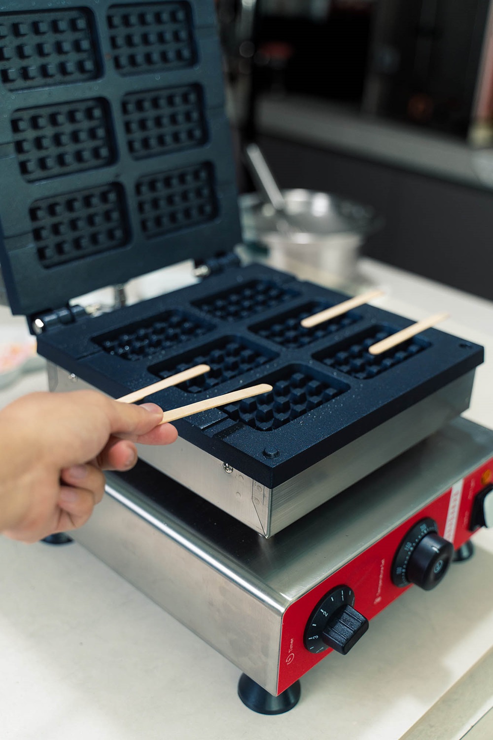 A hand places wooden sticks into a waffle maker with an open lid, preparing crispy popsicle waffles. The appliance, perfect for following your favorite popsicle waffle recipe, sits on a countertop.