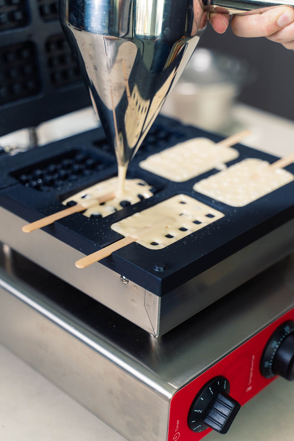 Batter is being poured from a metal dispenser into popsicle-shaped molds with sticks on a waffle maker, showing how to make crispy popsicle waffles using a popsicle waffle machine Malaysia.