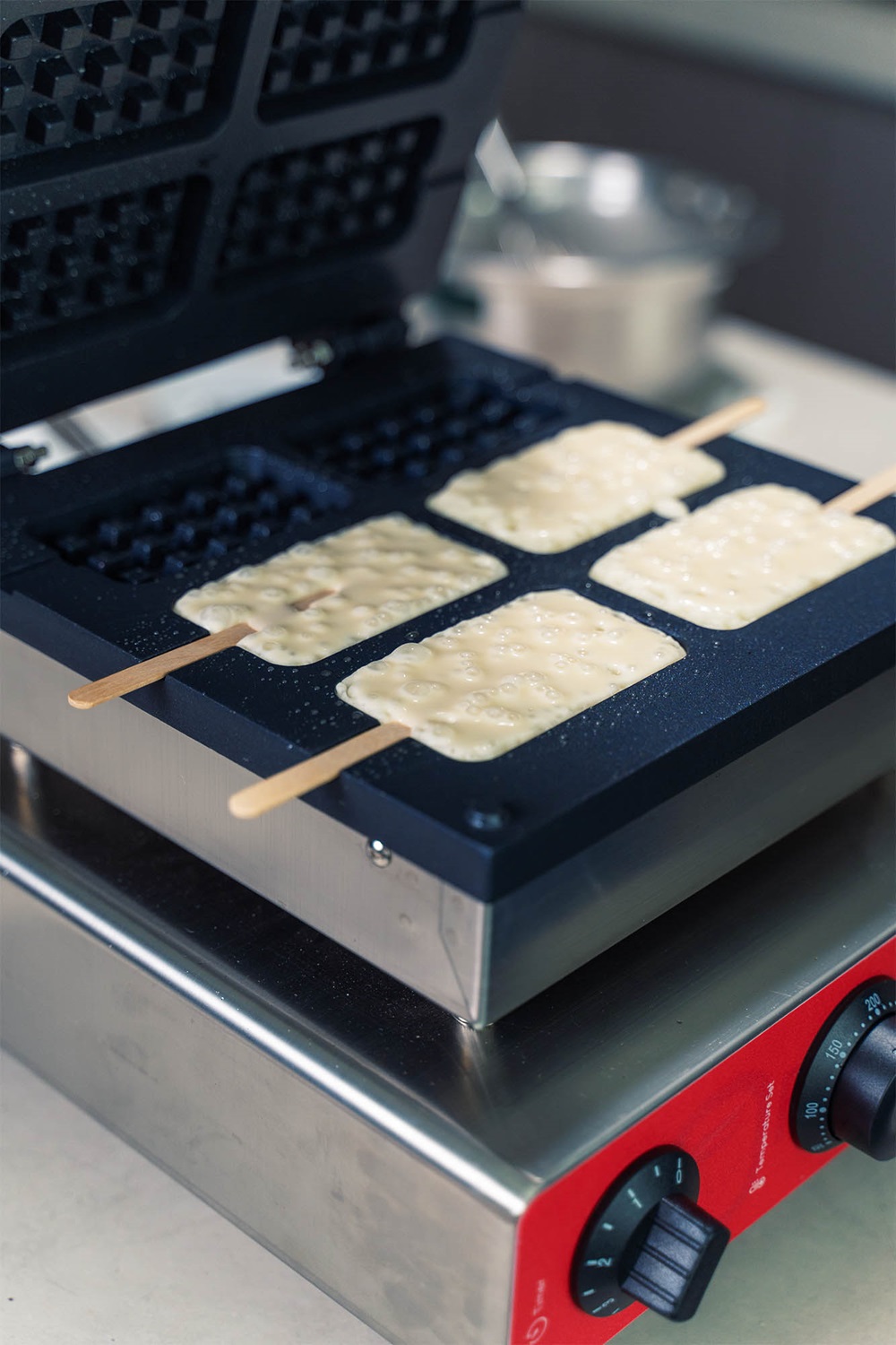 A popsicle waffle machine in Malaysia cooks four rectangular popsicle waffles on sticks, with the lid open and the batter visibly expanding.