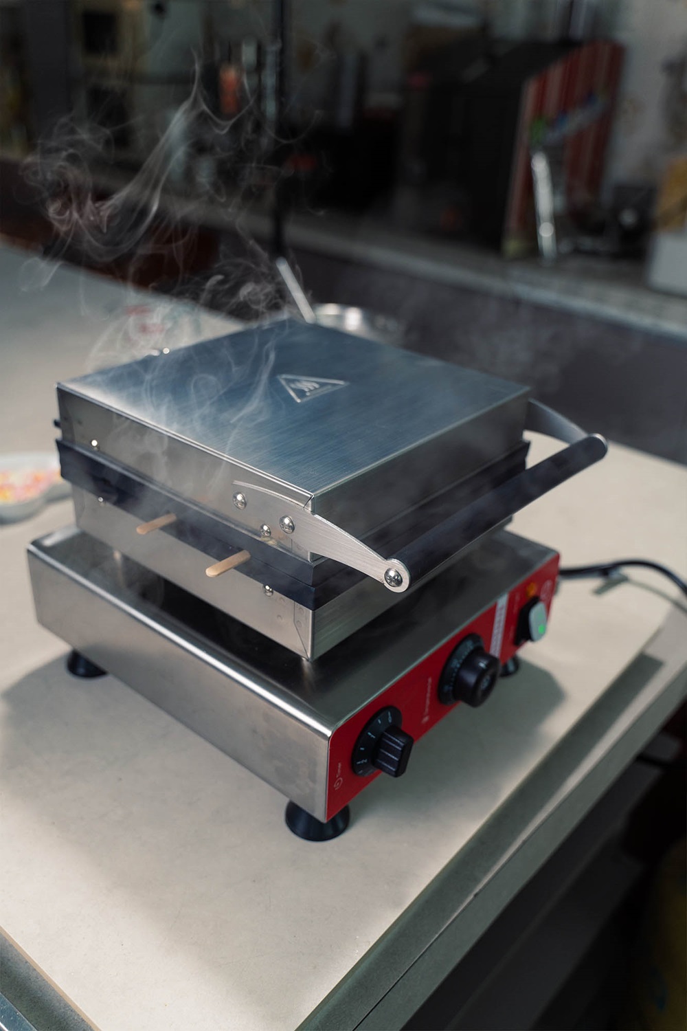 A closed stainless steel Popsicle waffle machine Malaysia sits on a countertop, plugged in and turned on, with steam rising, wooden handles, and control knobs visible.