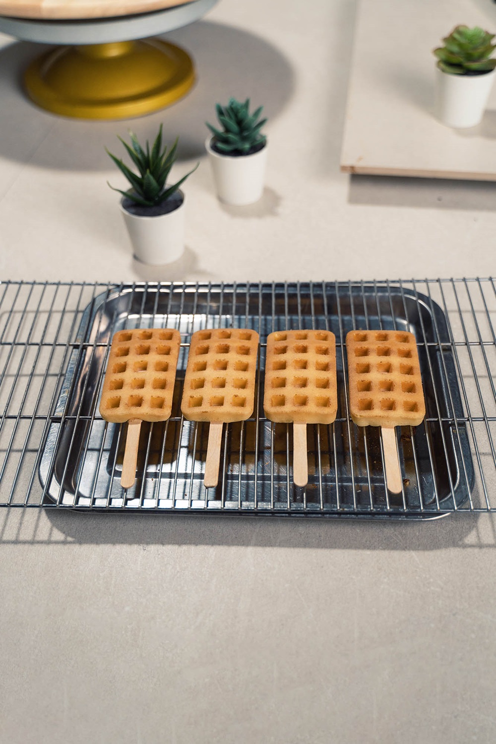 Four crispy popsicle waffles on wooden sticks are arranged in a row on a metal cooling rack, with three small potted succulents in the background—perfect inspiration for those wondering how to make waffle treats at home.