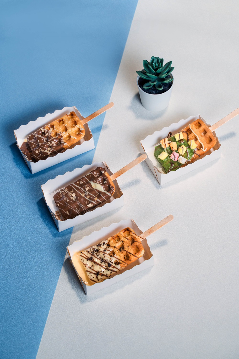 Four assorted Popsicle Waffles with various toppings are arranged in trays on a blue and white surface, with a small potted succulent in the background—a delightful inspiration for your next popsicle waffle recipe.