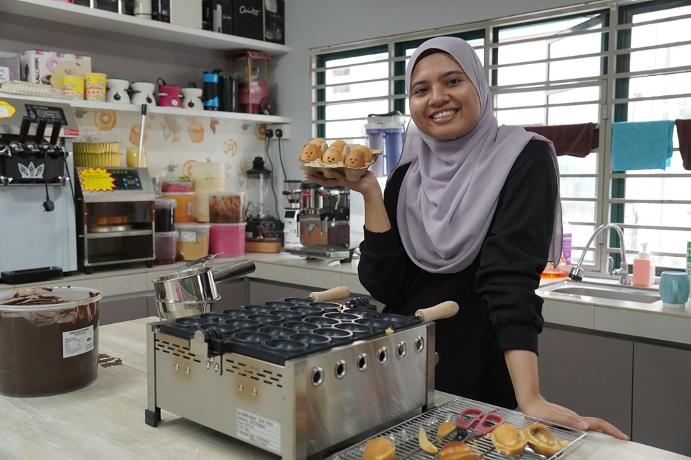 A woman in a hijab stands in a kitchen, smiling and holding cartoon-shaped baked goods made with an Egg Shape Waffle Maker, with baking equipment and supplies on the counter—perfect inspiration to Start Small Food Business dreams.