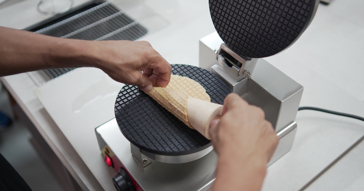 A person shapes a freshly baked waffle into a cone using a waffle cone machine on a countertop, similar to those found with leading ice cream cone maker brands in Malaysia.