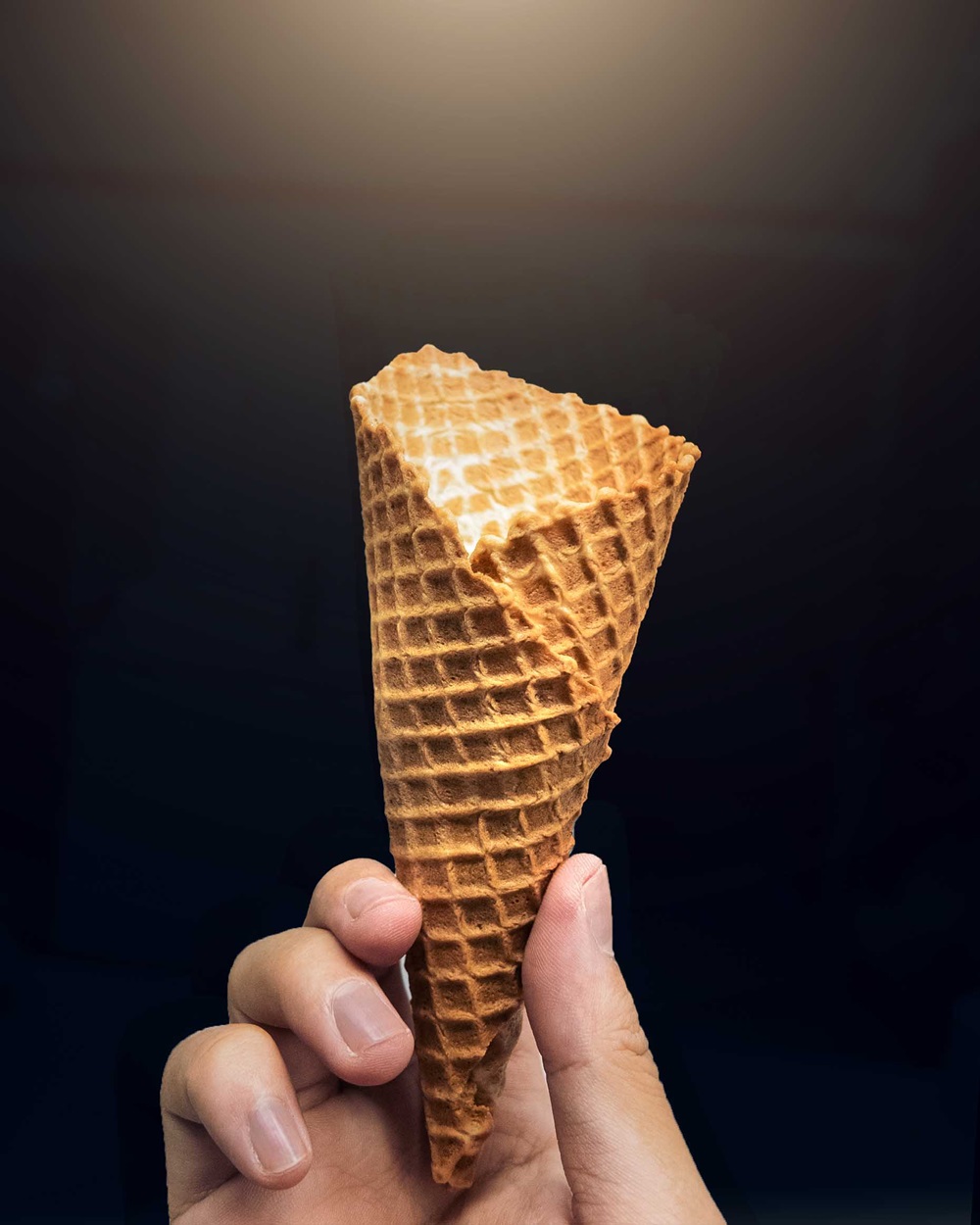 A hand holds an empty waffle cone made with a waffle cone machine against a dark background, softly lit from above.