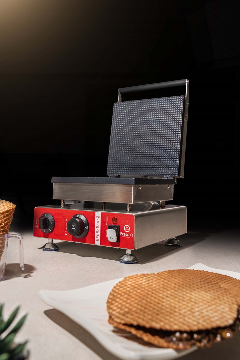 A commercial ice cream cone maker with a red control panel sits on the countertop, surrounded by waffle cones and a plate in the foreground.