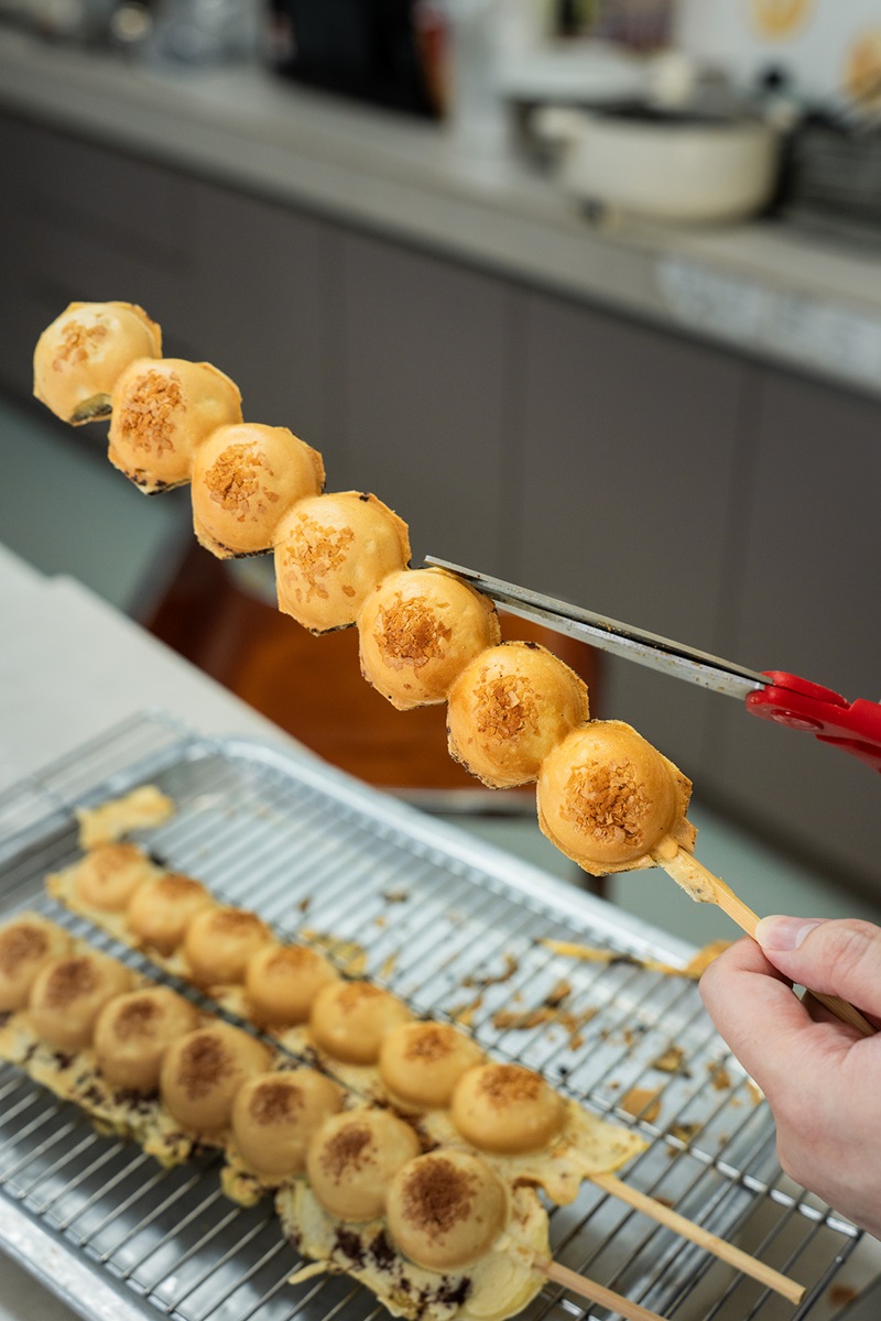 A person uses scissors to cut through a skewer holding round baked treats made from a waffle ball machine, with more skewers of waffle balls cooling on a wire rack below.