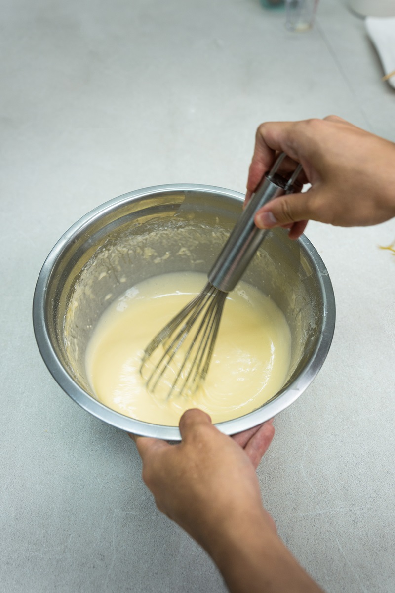 A person uses a metal whisk to mix a light-colored batter in a stainless steel bowl on a gray countertop while preparing a crispy waffle balls recipe.