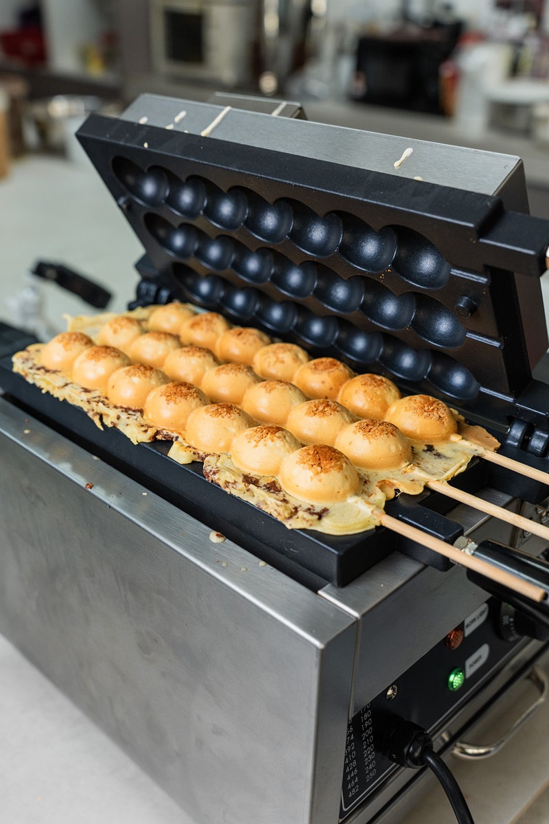 A bubble waffle maker with an open lid reveals golden brown waffle balls on skewers, perfect for learning how to make waffle ball treats at home.