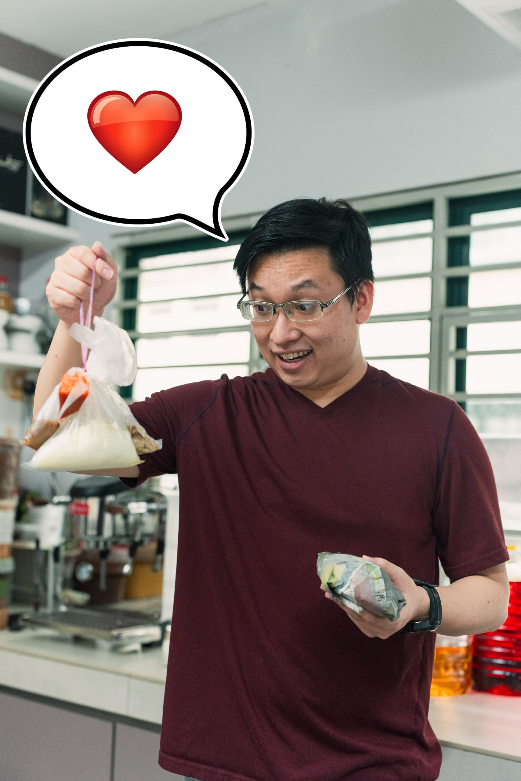 A man in a maroon shirt holds food sealed with a food sealer in plastic bags and a sandwich, smiling with a heart emoji in a speech bubble above him, standing in a kitchen.