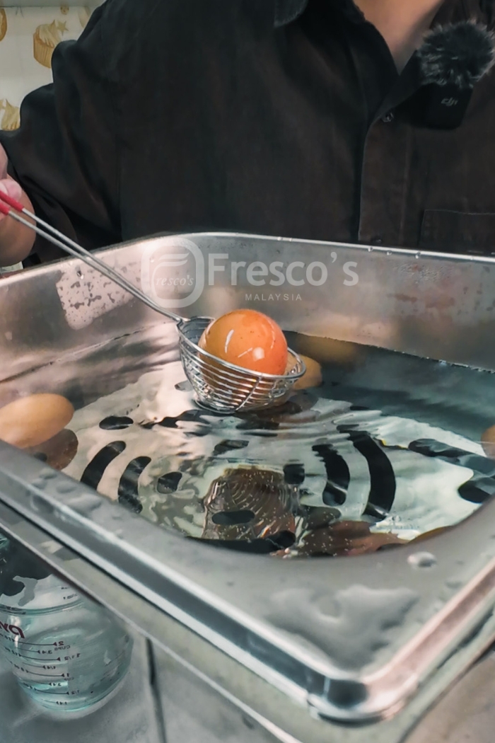 A person lifts a brown egg from a metal tray filled with water using a wire skimmer, preparing half-boiled eggs in a kitchen setting—an effortless method perfect for any egg cooker recipe.