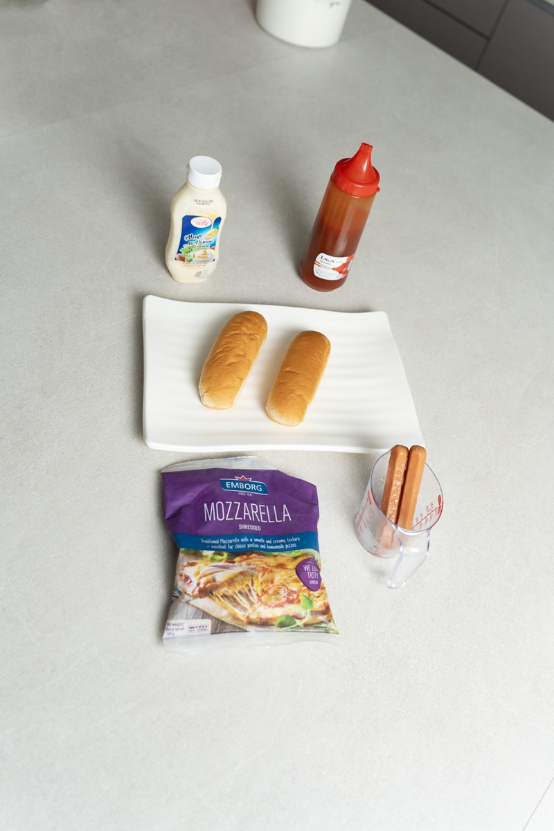 A countertop displays two hot dog buns on a plate, two steamed hotdogs in a cup, a bag of shredded mozzarella, ketchup, and a bottle of salad dressing.